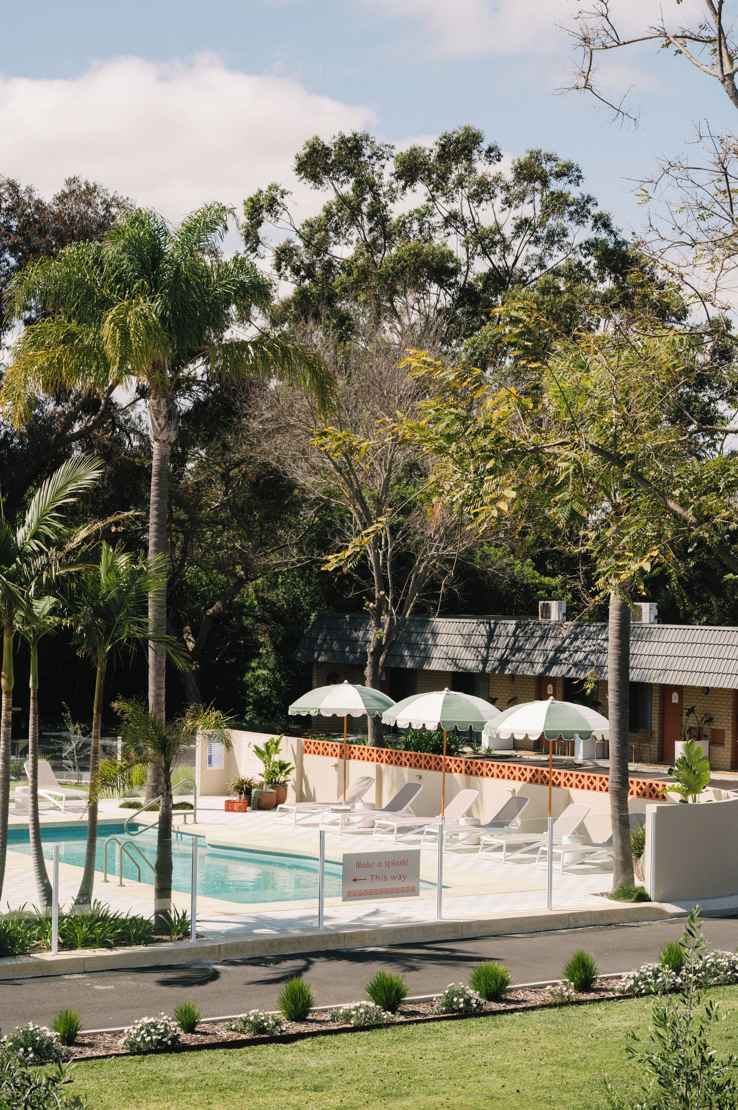 A pool area with lounge chairs and umbrellas, surrounded by palm trees and a building with a tiled roof.