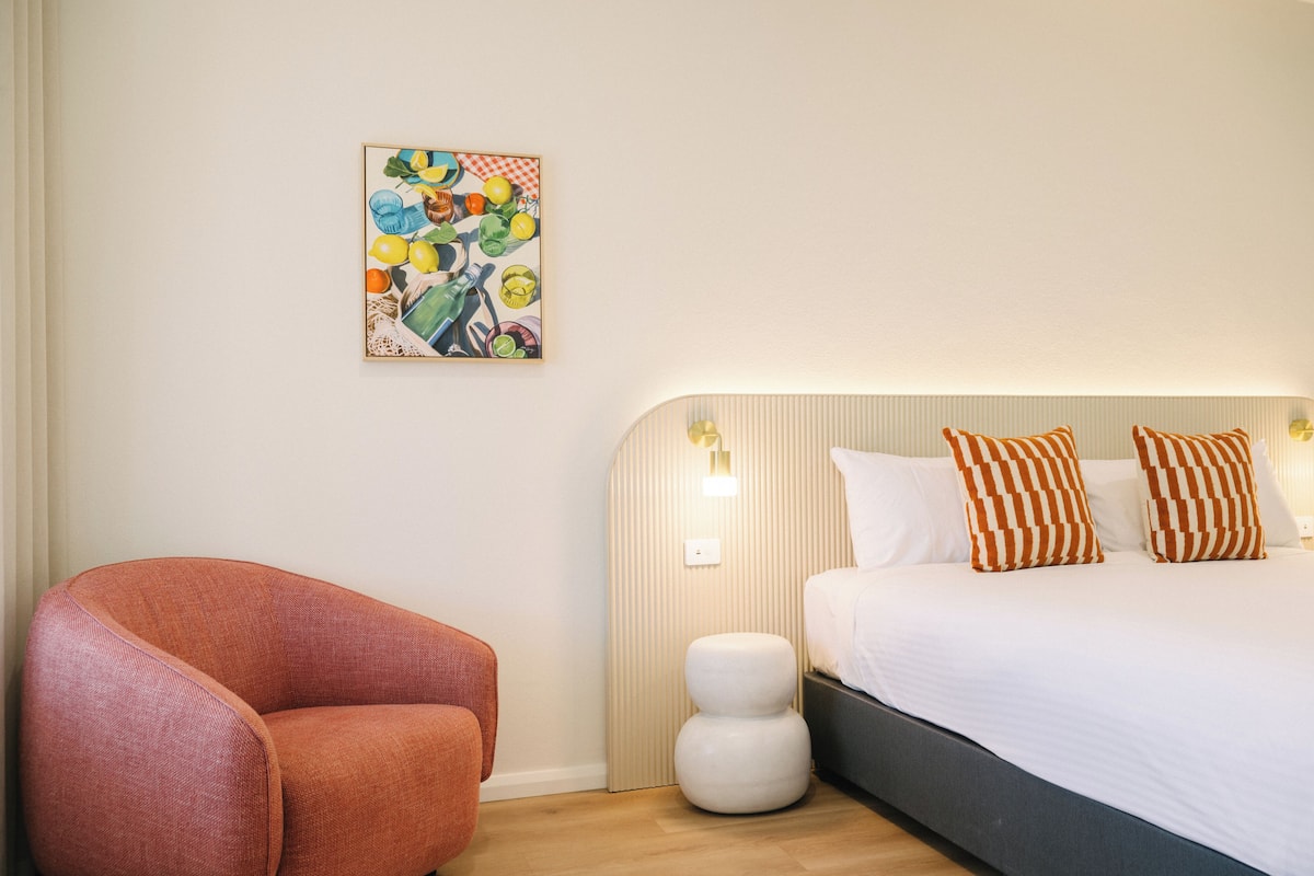 Una habitación de hotel moderna con una cama curvada, almohadones a rayas, un taburete blanco pequeño y un cuadro abstracto colorido en la pared.