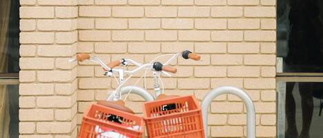 Una bicicleta verde clara con dos cestas naranjas sujetas al frente, estacionada frente a un muro de ladrillo con una ventana y una cámara de seguridad.
