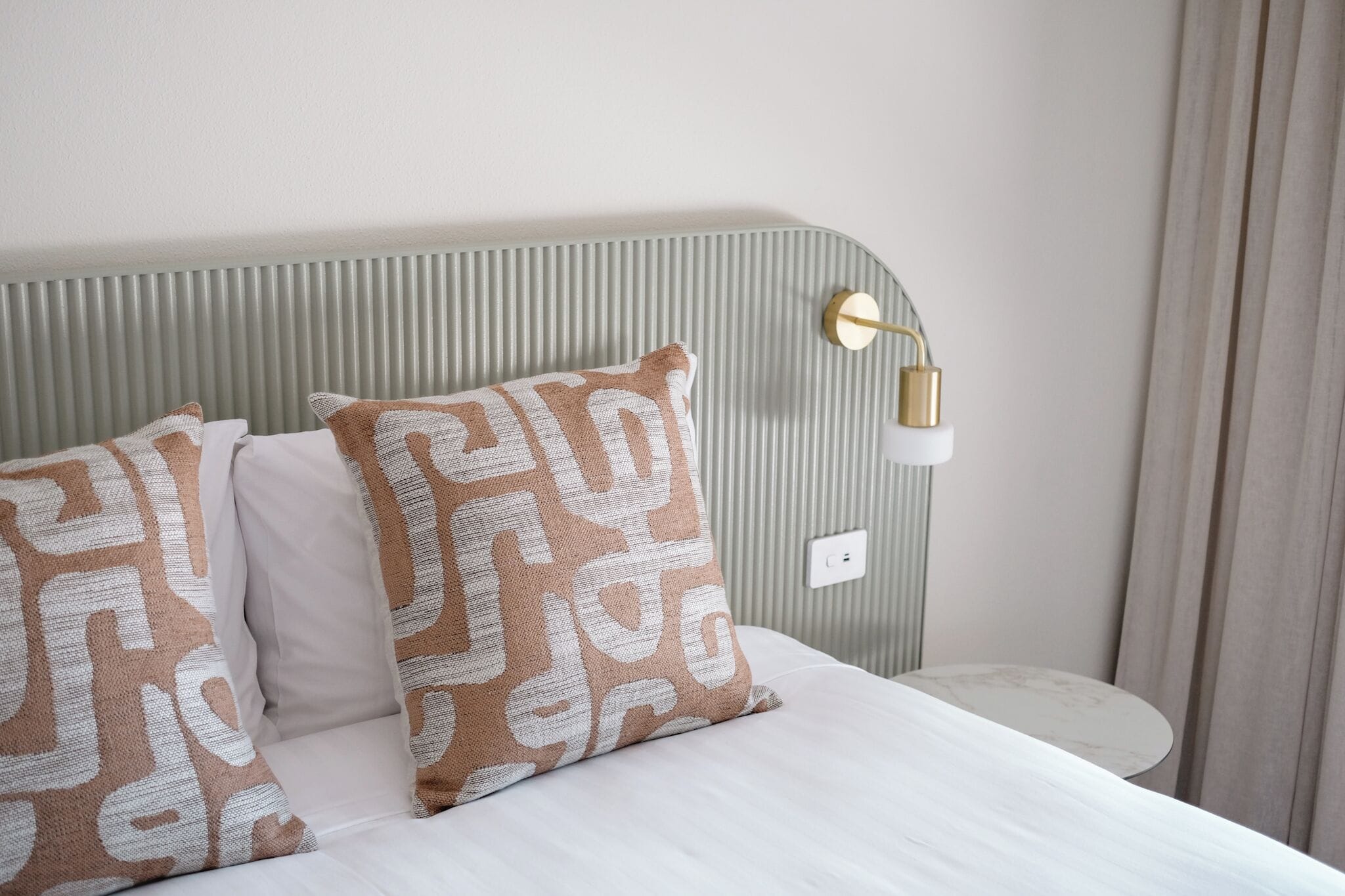 Una cama bien hecha con almohadones estampados, una mesa redonda y una lámpara de pared.