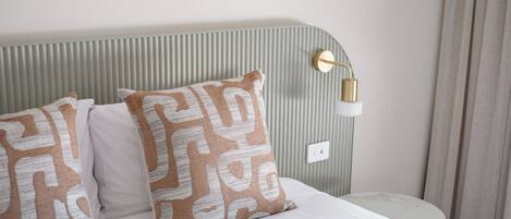 A neatly made bed with patterned pillows, a round table, and a wall-mounted lamp.