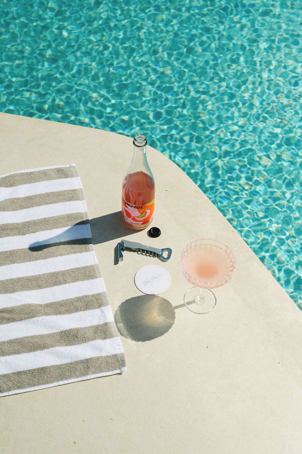Una toalla de playa, una botella de jugo de naranja, un abrelatas, un vaso con una bebida rosada y un vaso de Tim Hortons sobre una superficie de concreto al lado de una piscina.