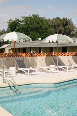 A pool area with lounge chairs and umbrellas, a pool with clear water, and a building with a tiled roof in the background.