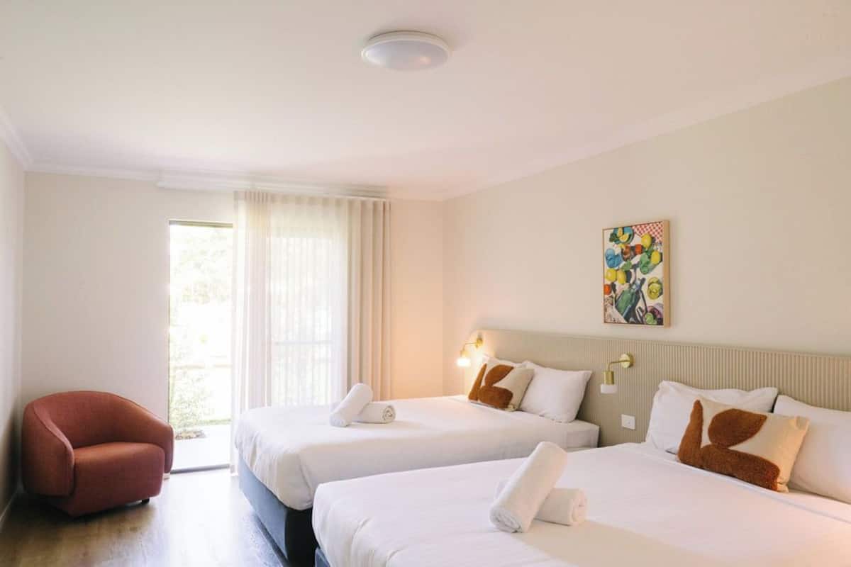 Habitación de hotel con dos camas, un sillón rojo, un ventanal grande con cortinas, una obra de arte colgada en la pared y una lámpara de techo.