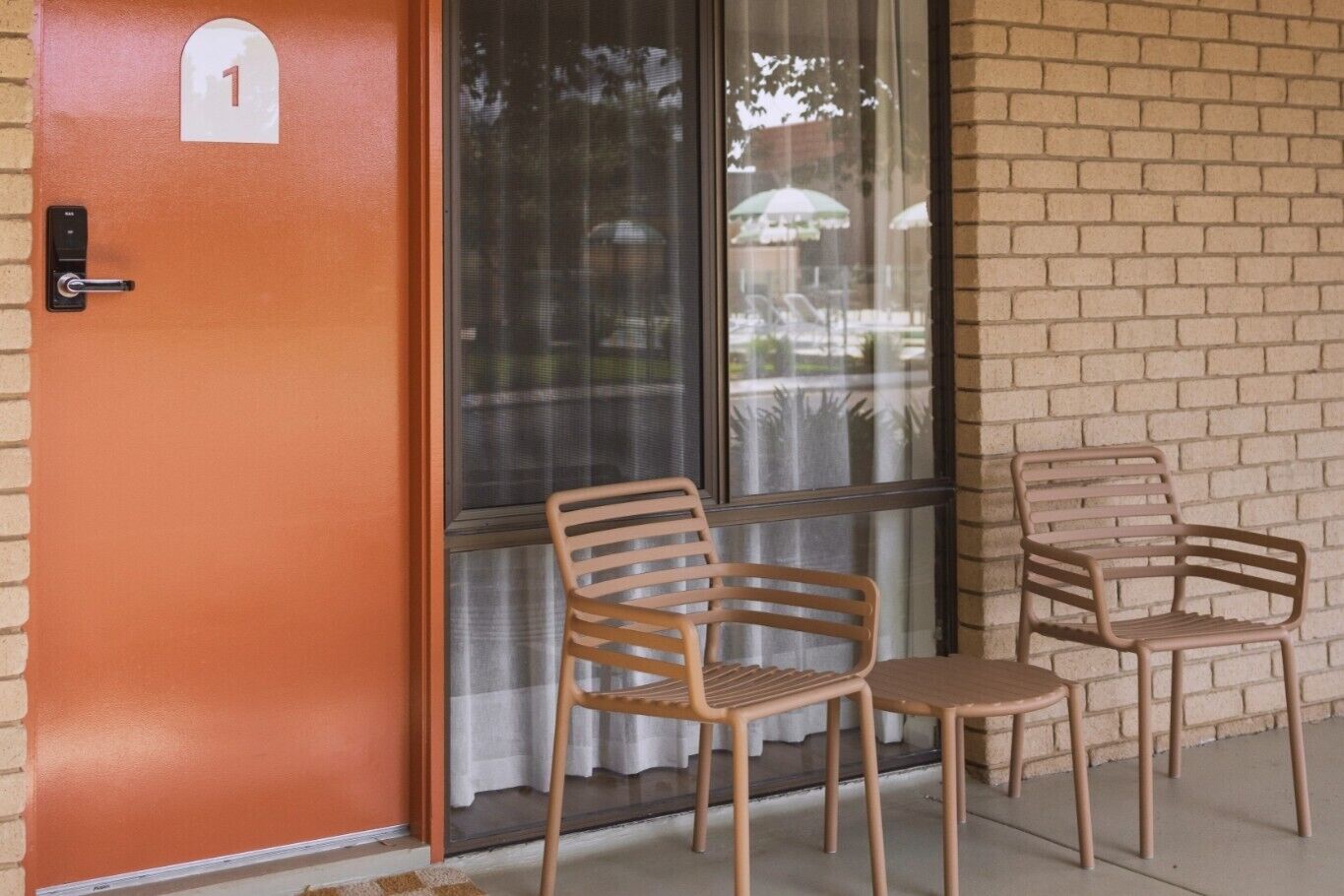 Una puerta con un letrero del número 1, dos sillas de madera y un ventanal con vistas a sombrillas exteriores.
