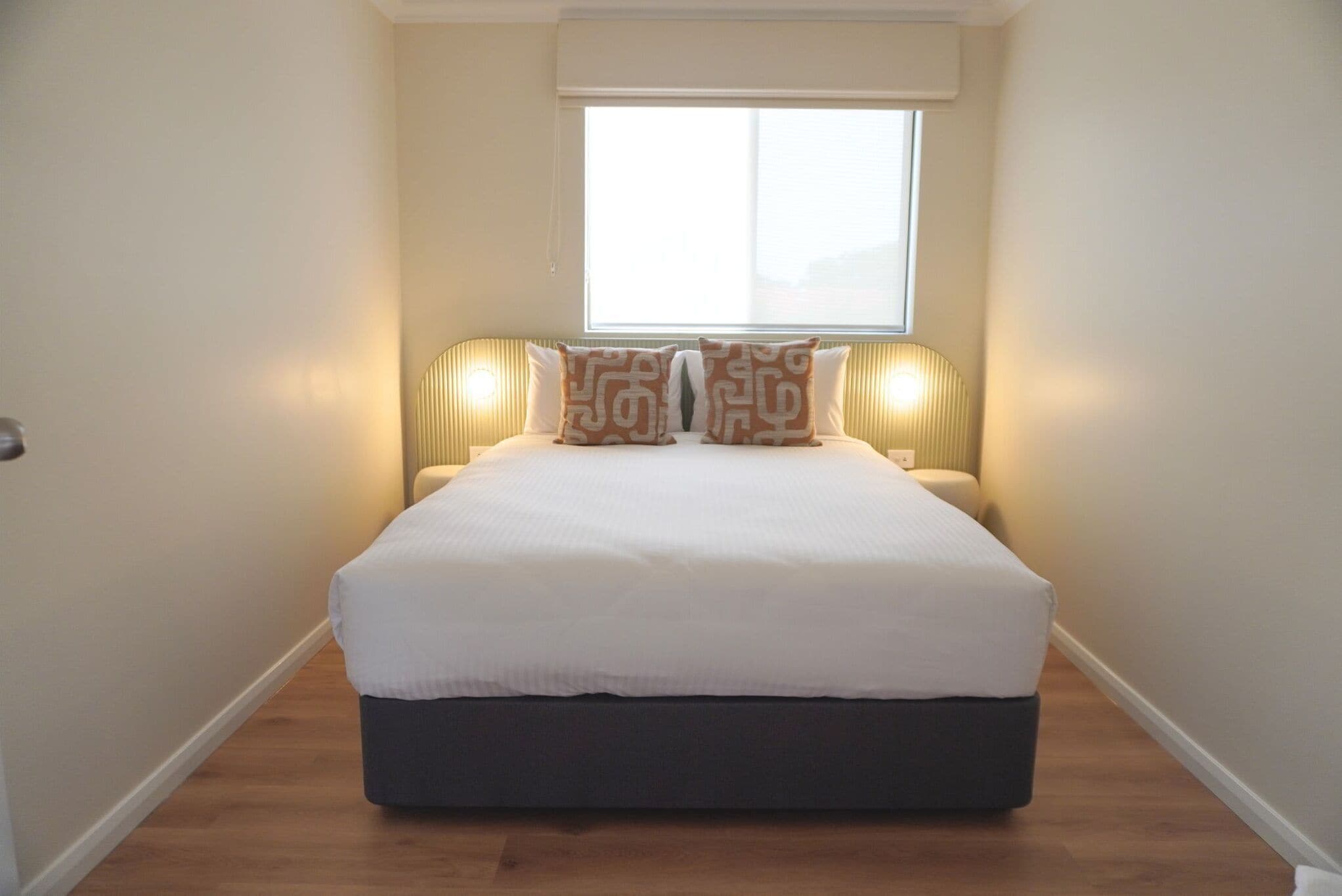 A narrow, well-lit bedroom with a large bed, two bedside lamps, and a window with blinds.