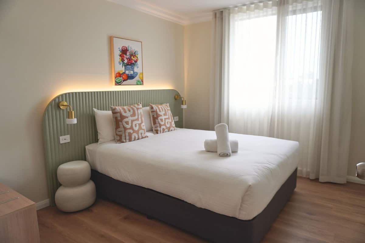 Una cama bien tendida con almohadas, una lámpara de noche y una obra de arte en la pared.