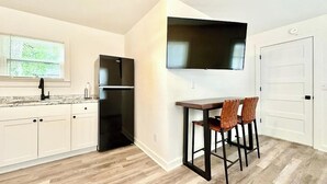 Una cocina con refrigerador, una mesa de comedor con sillas, un televisor montado en la pared y una ventana con persianas.