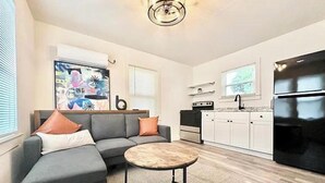 Una sala de estar moderna con un sofá modular, una mesa de centro redonda y una cocina con armarios blancos y un refrigerador negro.