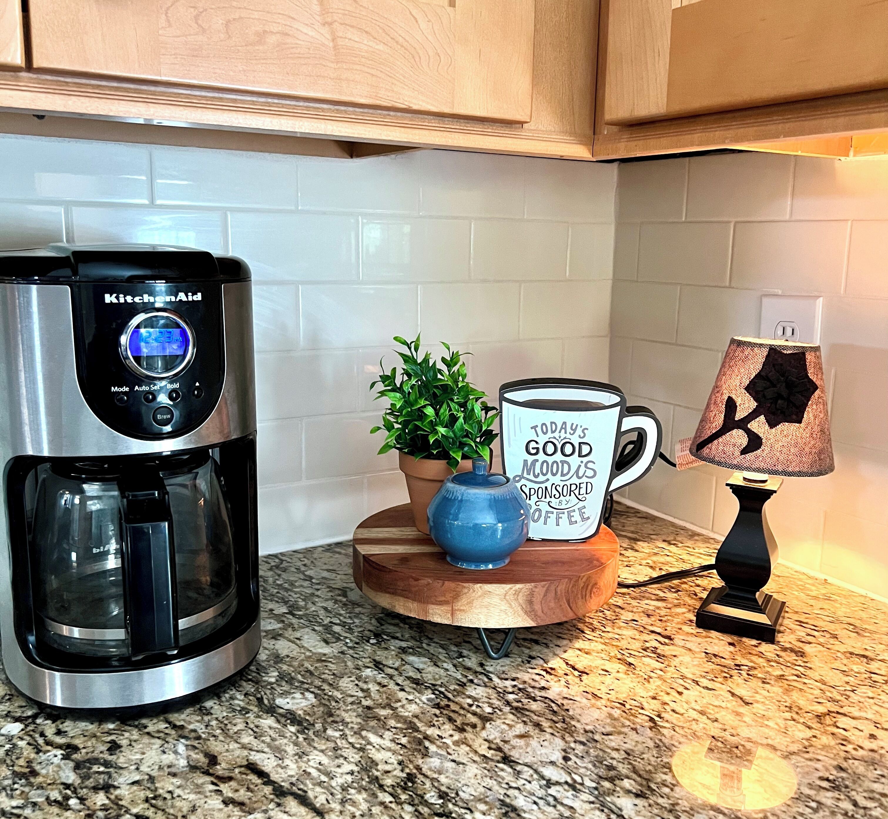 Une cuisine équipée d’une machine à café, d’une plante en pot, d’une tasse et d’une lampe posée sur un support en bois.