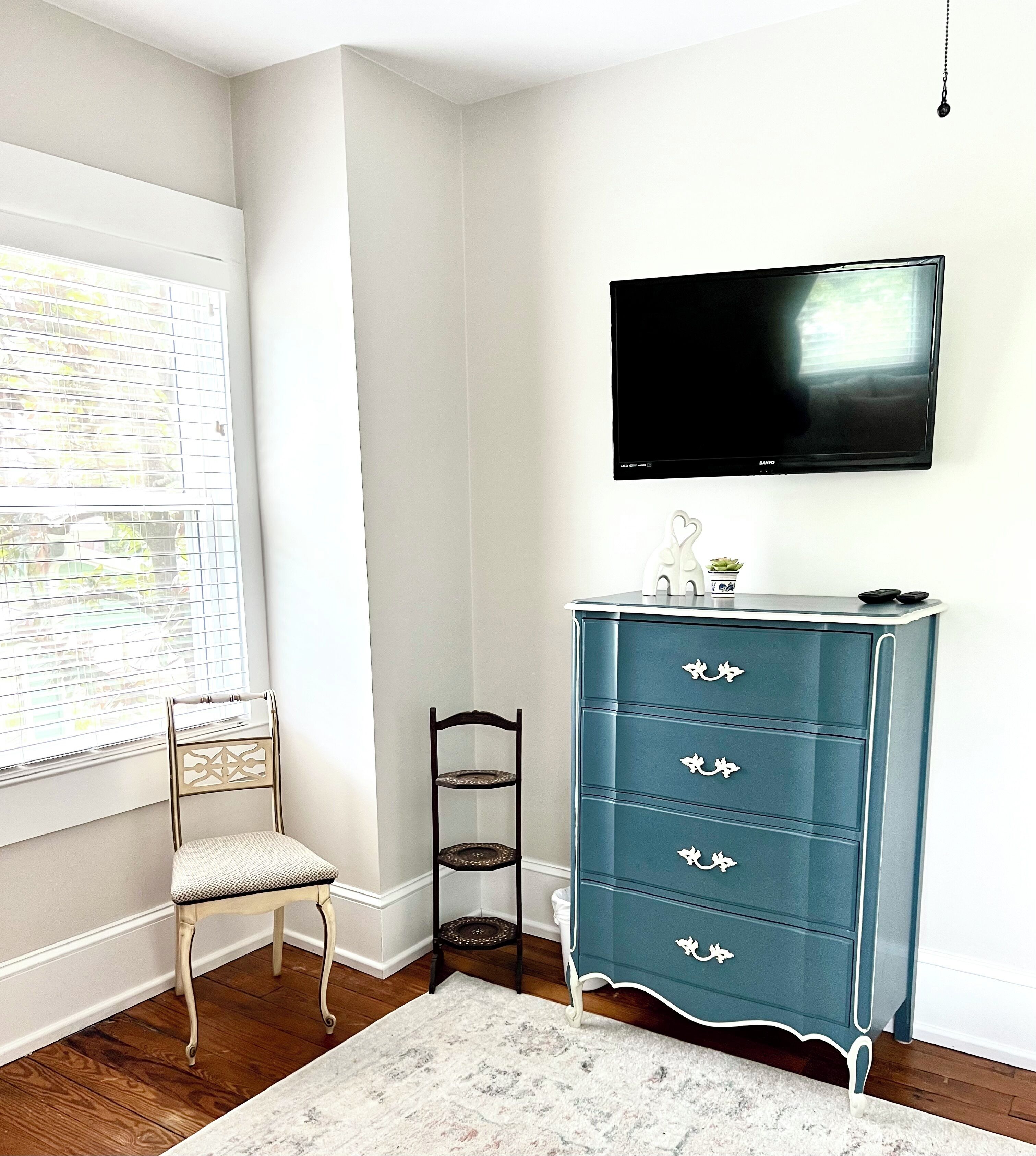 A room with a wooden floor, a white chair, a teal dresser with drawers, a television mounted on the wall, and a window with blinds.