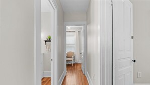 Un pasillo con piso de madera, paredes blancas y una puerta que da a una habitación con una silla y una mesita con una planta.