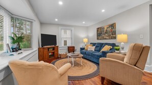 Una sala de estar con un sofá azul, sillones beiges, una mesa de centro de madera y un televisor sobre un mueble bajo.