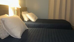 A hotel room with two beds, a nightstand, a lamp, and a window with curtains.
