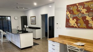 A modern kitchen with a black countertop, white cabinets, and a wooden dining table. There is a clock on the wall and a painting above the cabinets. - 1BR (sleeps 3) - private, modern granny flat with large shaded pool (Parap)