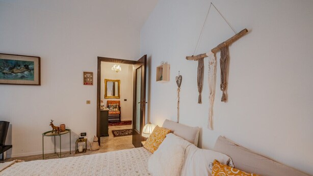 Une chambre à coucher avec un lit, des oreillers, une petite table, une chaise, un miroir et des décorations murales en macramé.