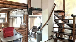 A rustic interior with exposed wooden beams, a staircase, a seating area with a chair and a table, and a window with curtains.