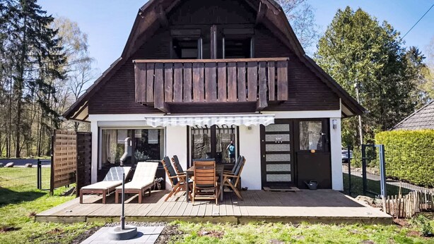 A wooden chalet with a balcony, a patio with outdoor seating, and a view of the surrounding forest.
