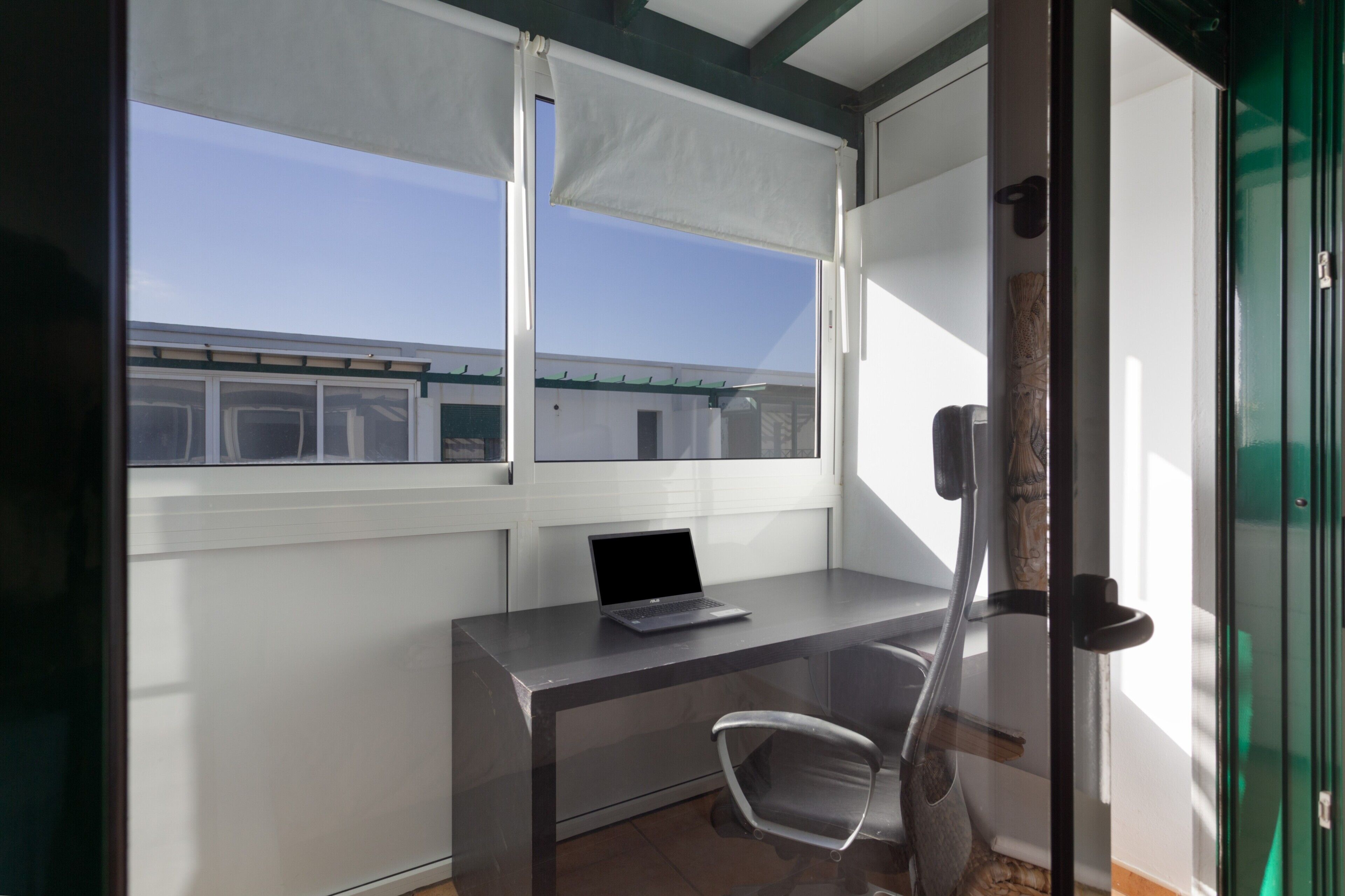 Een kamer met een bureau, een stoel, een laptop, een raam met zonwering en een deur met een ruit.