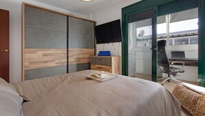Un dormitorio con cama, escritorio, silla, armario y puerta corrediza de vidrio a un balcón.