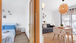 A room with a bed, a nightstand, a bedside table, a sofa, a dining table with chairs, a hanging light fixture, and a window with curtains.
