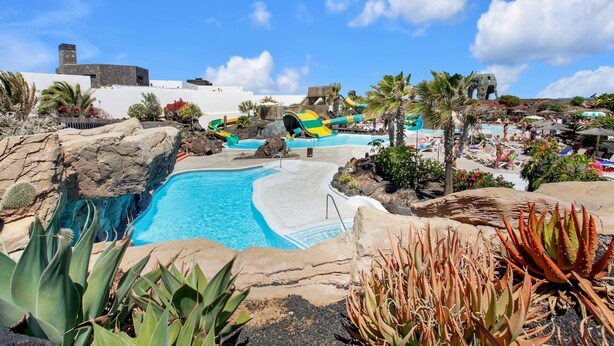 A pool area with water slides, palm trees, and a rocky landscape.