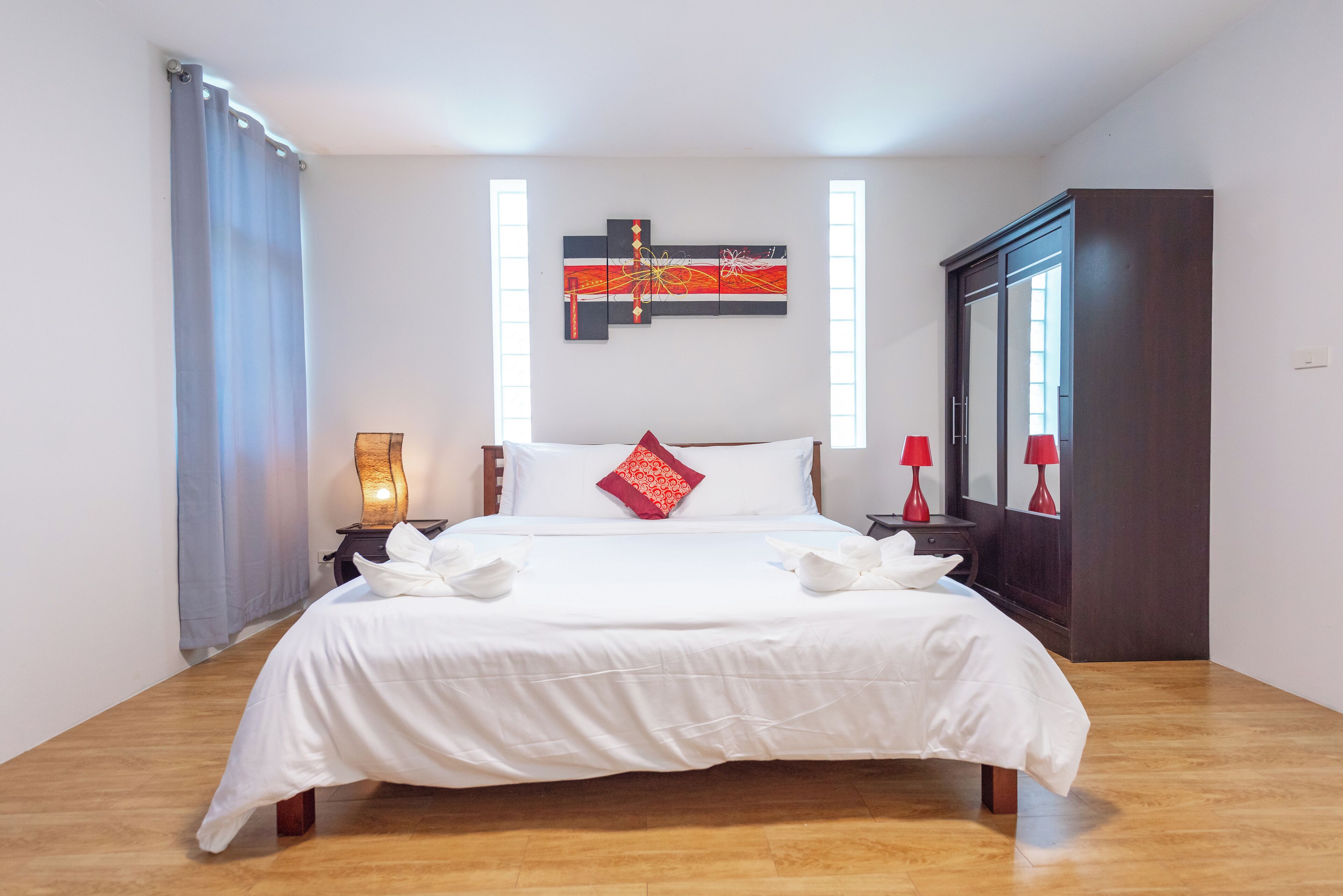 A neatly made bed with white linens and a red accent pillow, a bedside table with a lamp, and a large mirror on the wall.
