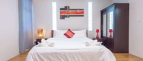 Un lit bien fait, avec du linge de lit blanc et un coussin rouge, une table de chevet avec une lampe, et un grand miroir au mur.