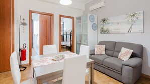 Un espace repas comprenant une table, des chaises et un canapé. Un tableau est accroché au mur, ainsi qu’un extincteur, et une porte donne accès à une autre pièce.