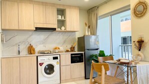 A compact kitchen with wooden cabinets, a washing machine, a refrigerator, and a dining area with a glass table and chair. - Amazing new 1-bedroom apartment, fabulous Lamai Beach,  swimming pool, fitness (Koh Samui)