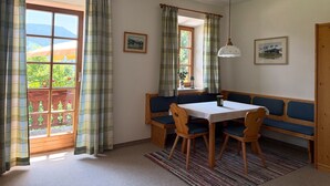 Ein gemütlicher Essbereich mit Tisch und Bänken, großen Fenstern mit Vorhängen und Blick ins Grüne.