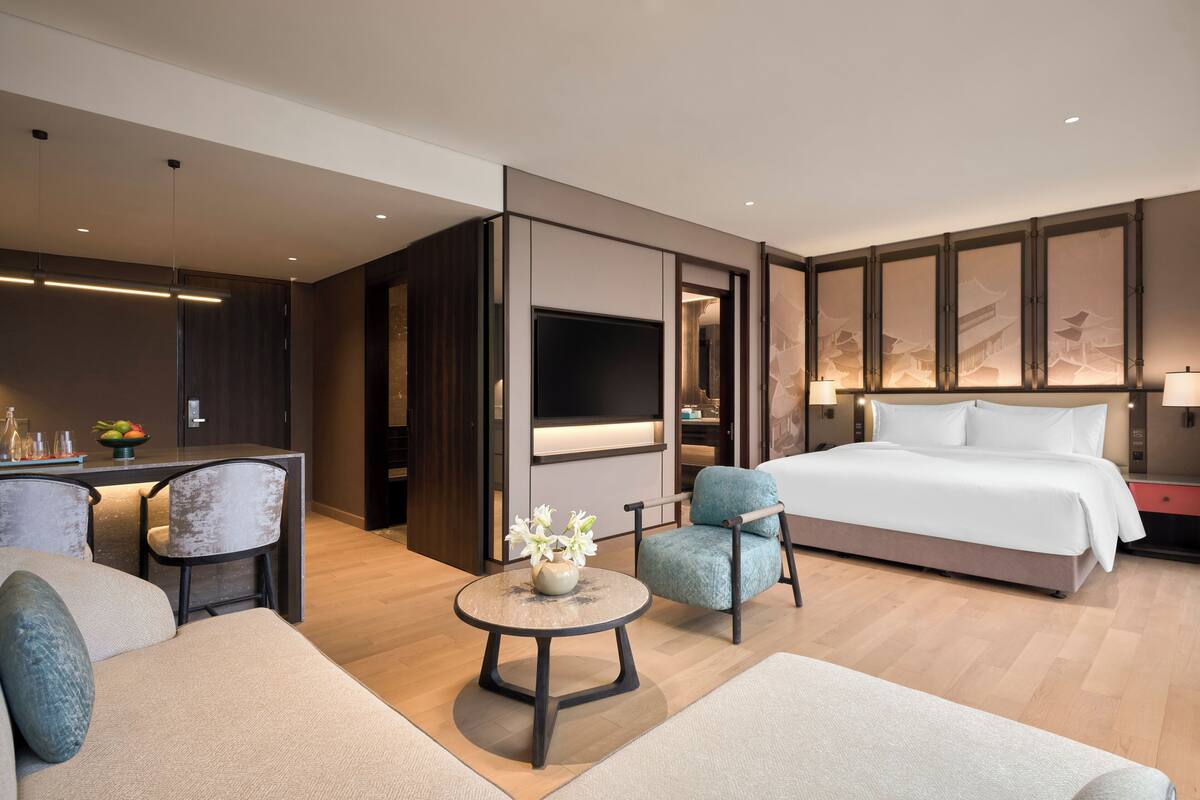 Une chambre d’hôtel moderne avec un grand lit, un canapé, une petite table ronde agrémentée d’un vase, un bureau avec une télévision et un mur orné d’œuvres d’art encadrées.