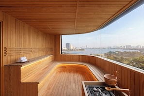 A sauna room with wooden walls and a curved bench, a view of a city skyline, and a window overlooking a body of water. - Dusit Le Palais Tu Hoa Hanoi (Hanoi)