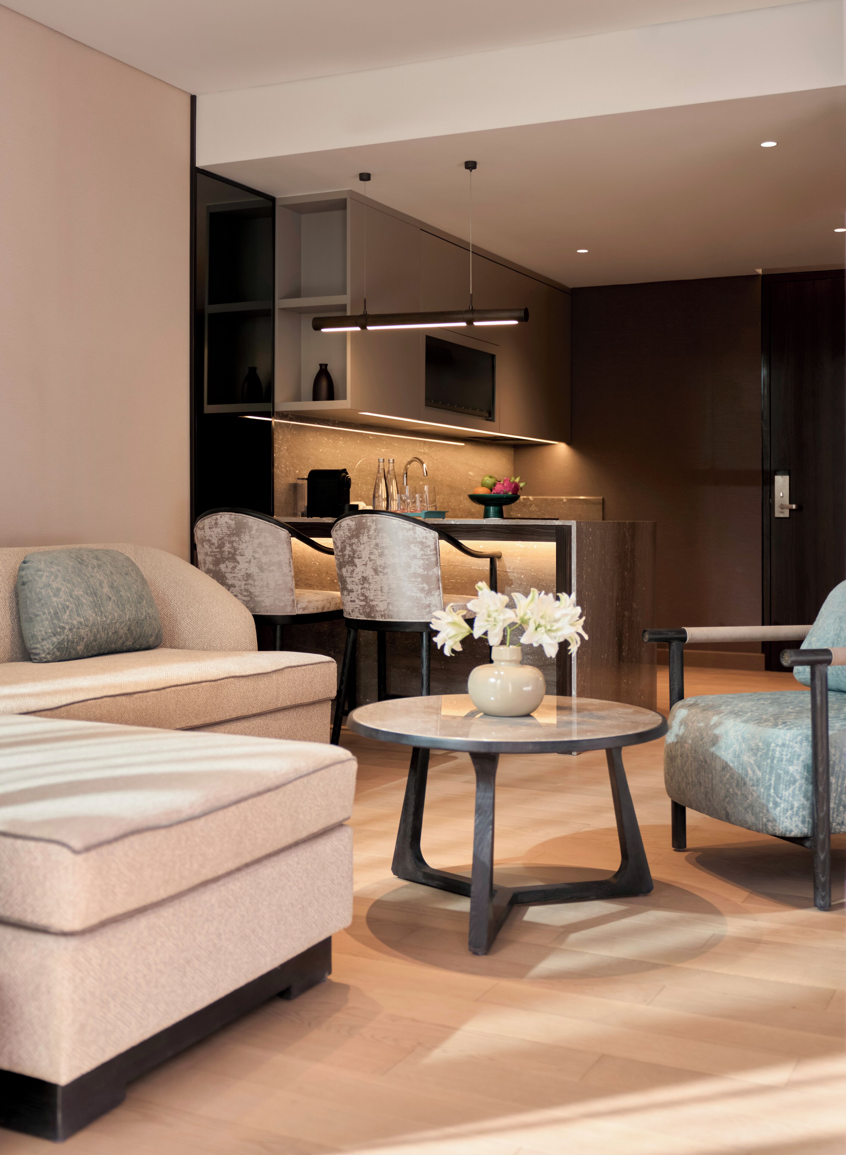 A modern living room with a sofa, a round table with a vase of flowers, and a kitchen area in the background.