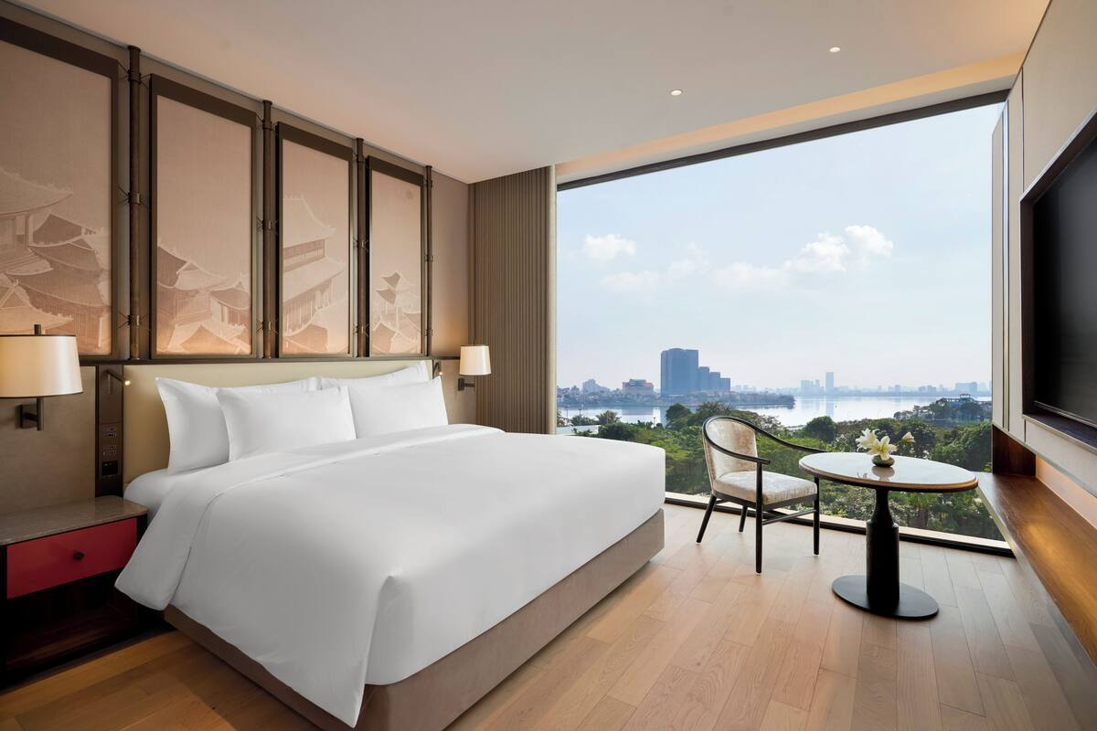 Habitación de hotel moderna con una cama grande, una mesita con un jarrón y vistas a un paisaje urbano a través de un ventanal amplio.