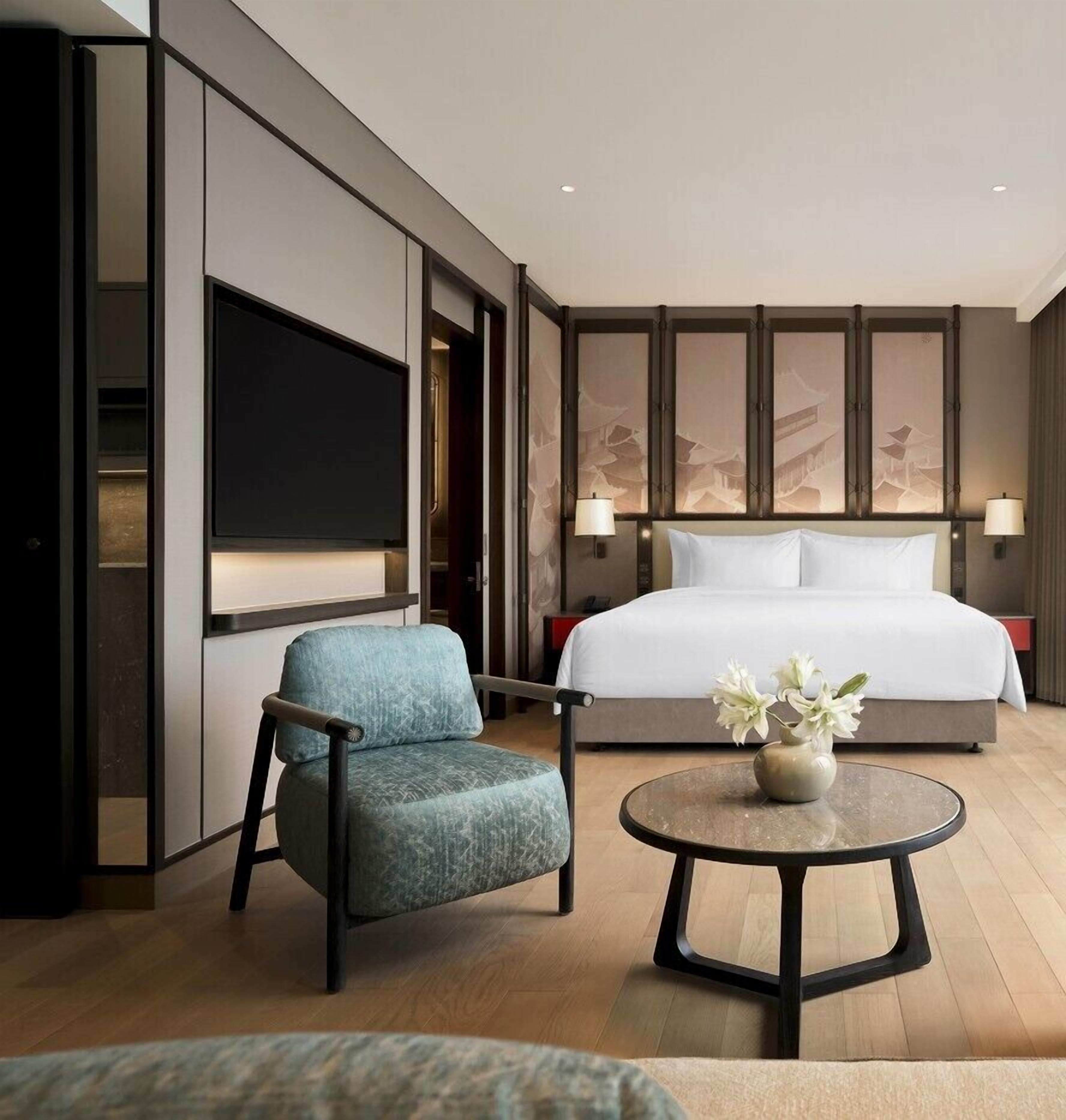 A modern hotel room with a large bed, a flat-screen TV, a blue armchair, a round coffee table with a vase of flowers, and traditional wall art.
