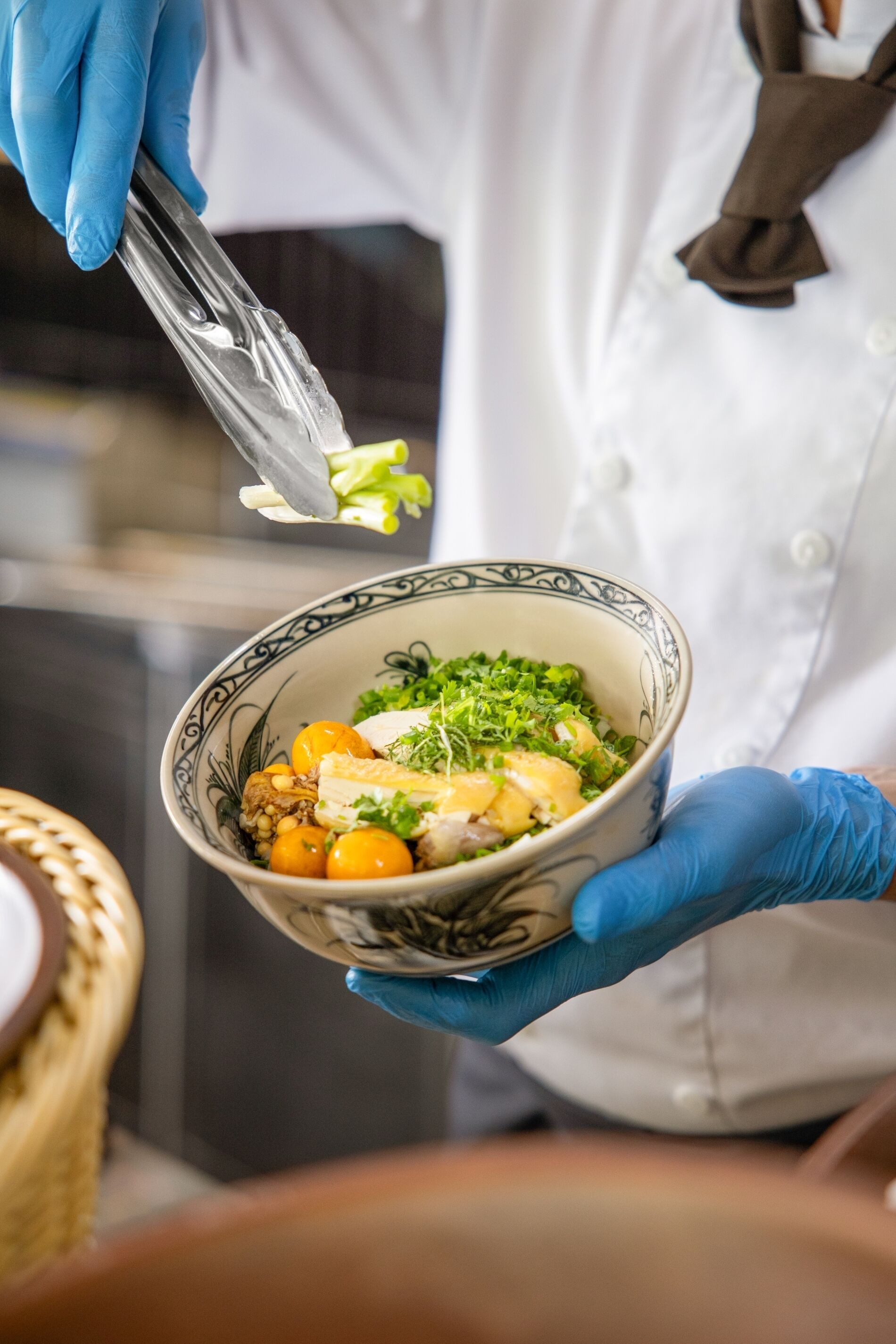 A person in a white chef's coat and blue gloves holding a bowl of food with tongs.
