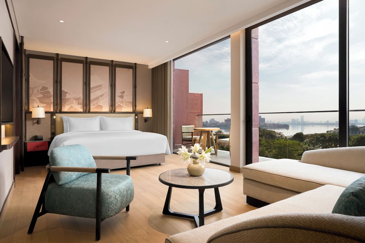 Une chambre d’hôtel moderne dotée d’un grand lit, d’un coin salon avec un canapé et un fauteuil, d’une petite table avec un vase et d’un balcon offrant une vue sur la ville.