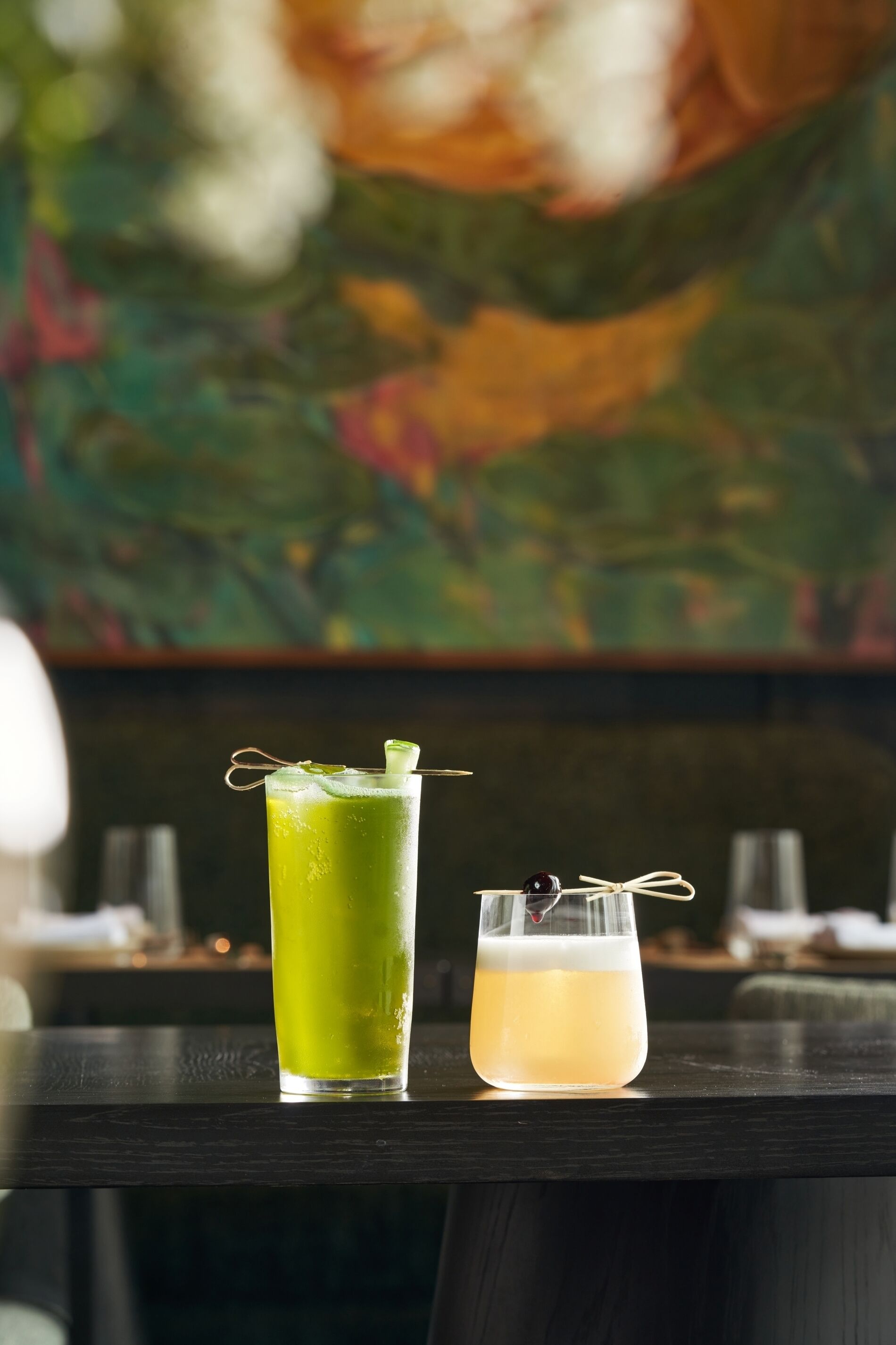 Two drinks on a table with a painting in the background.