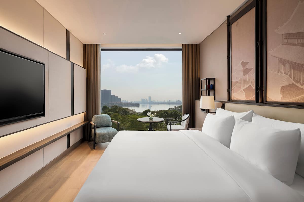 Une chambre d’hôtel avec un grand lit, un téléviseur à écran plat, une petite table avec un vase, et une vue sur le paysage urbain et l’eau.