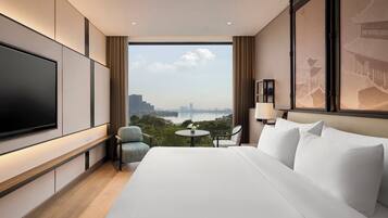 Een hotelkamer met een groot bed, een flatscreen televisie, een kleine tafel met een vaas en uitzicht op een stadsbeeld en water.
