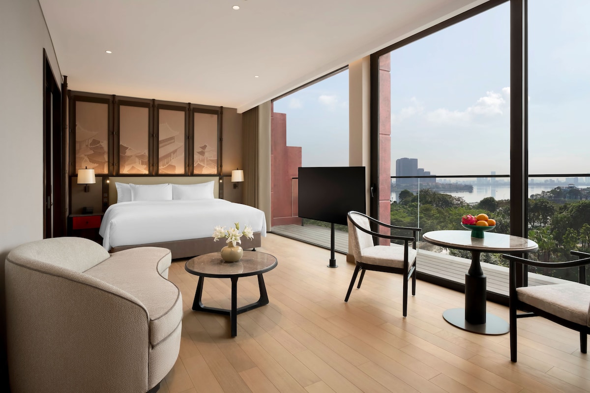 Une chambre d’hôtel moderne avec un grand lit, un canapé, une petite table avec un vase et un balcon offrant une vue sur la ville et l’eau.