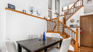 A dining area with a wooden table and white upholstered chairs, a staircase with wooden railings, and a wall-mounted shelf with framed artwork and a clock. - 2 Bedroom, beautiful end unit (Galena)