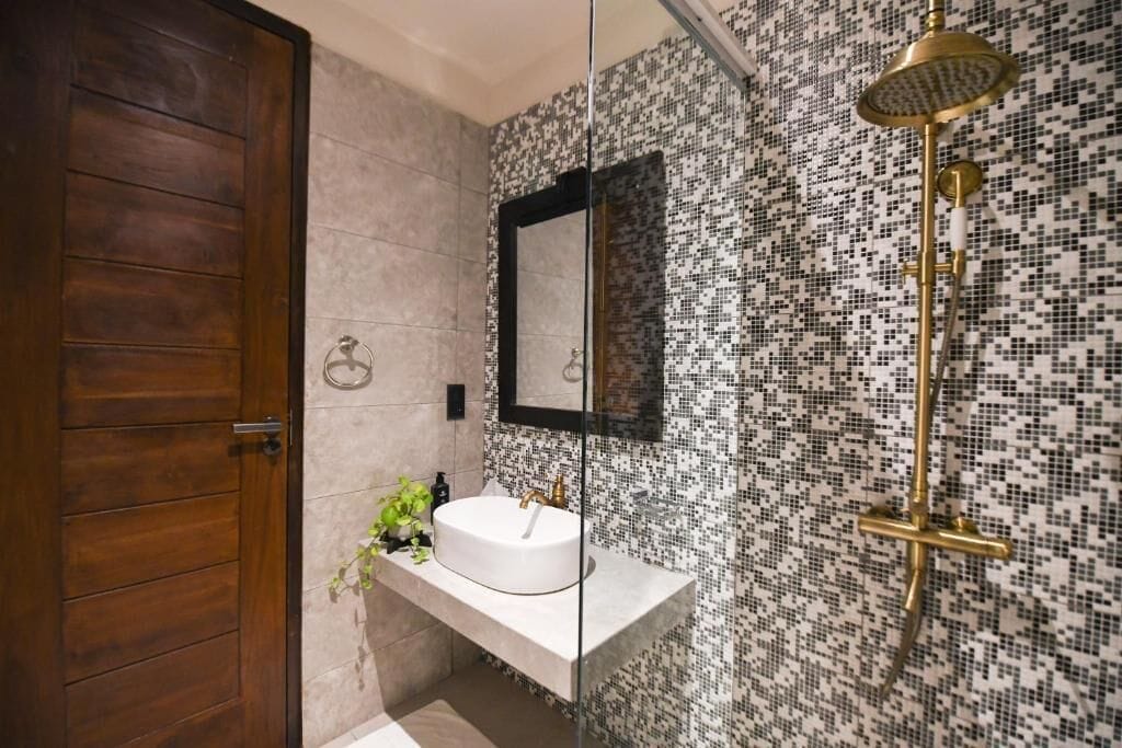 A modern bathroom with a wooden door, a white sink, a mirror, and a mosaic-patterned shower wall.