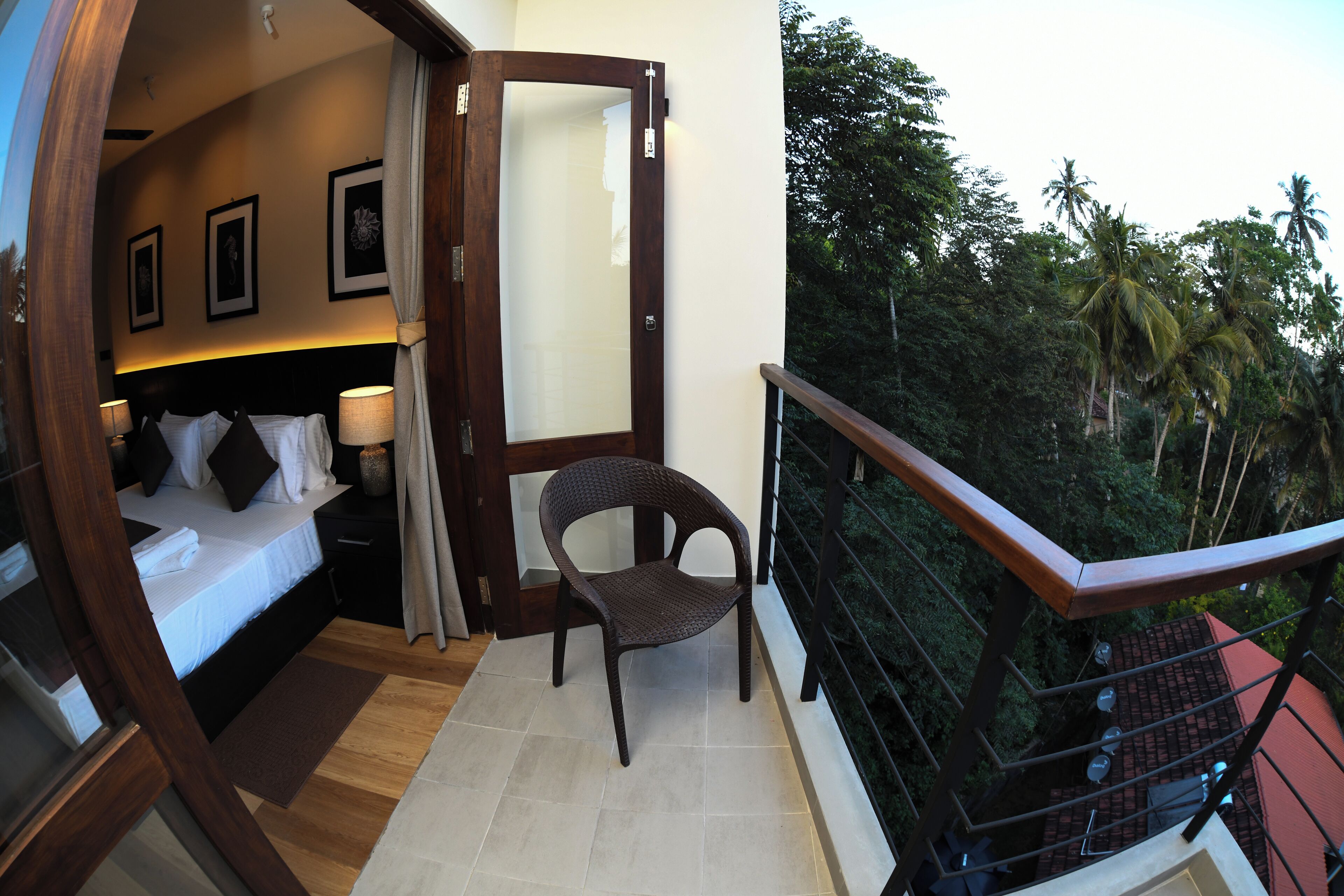 A balcony with a wooden railing, a bed with white linens, a wicker chair, and framed pictures on the wall.