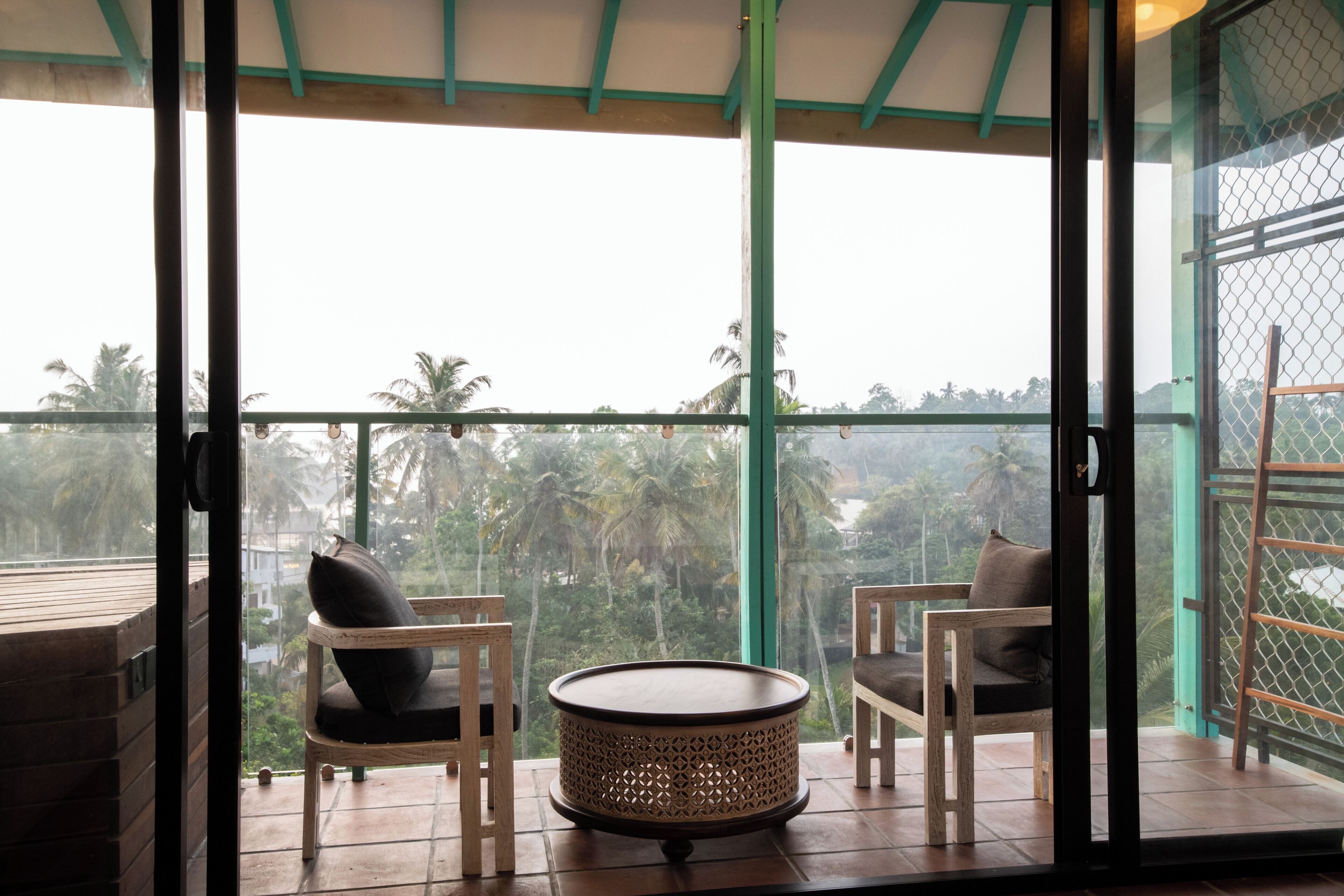 A balcony with two wooden chairs and a woven table, overlooking a lush green landscape with palm trees.