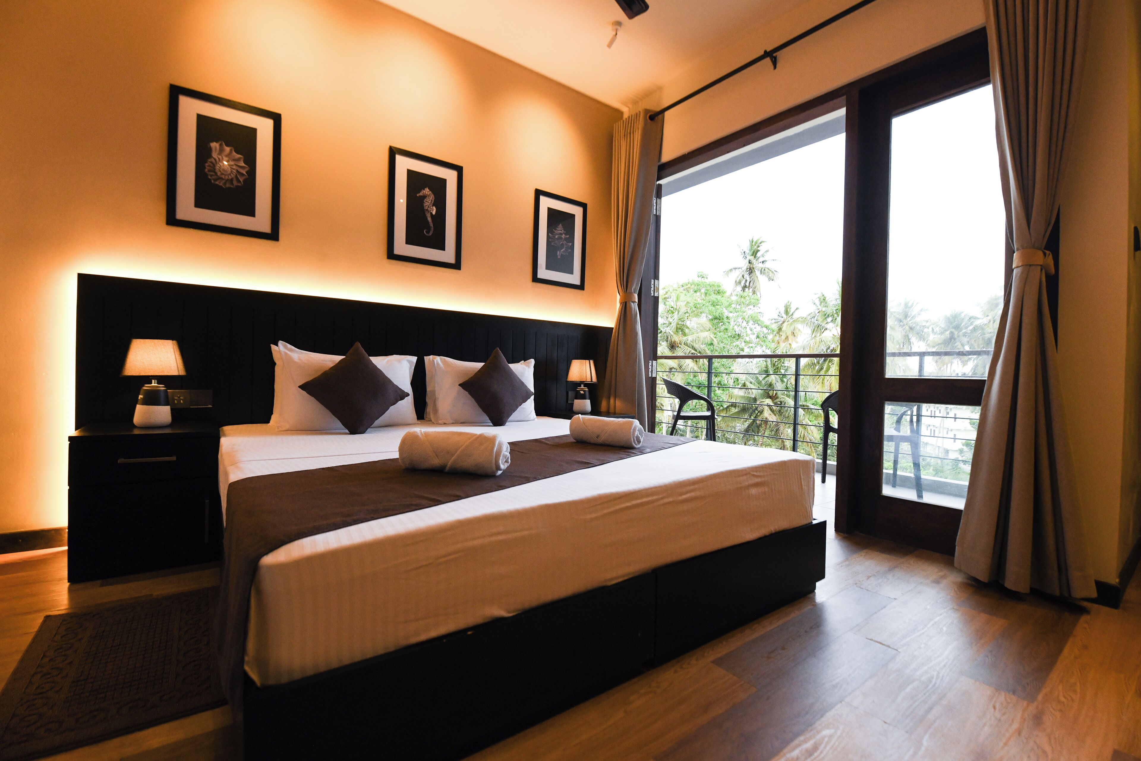 Una habitación de hotel moderna con una cama grande, dos mesitas de noche con lámparas, una obra de arte enmarcada en la pared y un balcón con vistas a zonas verdes.