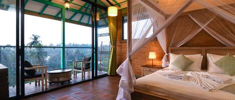 A bedroom with a bed, a nightstand, a lamp, a chair, a small round table, and a view of the outdoors through large windows.
