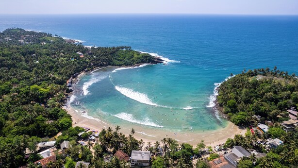 Une vue aérienne d’une plage aux eaux turquoise et limpides, bordée d’une végétation luxuriante et ponctuée de quelques bâtiments.