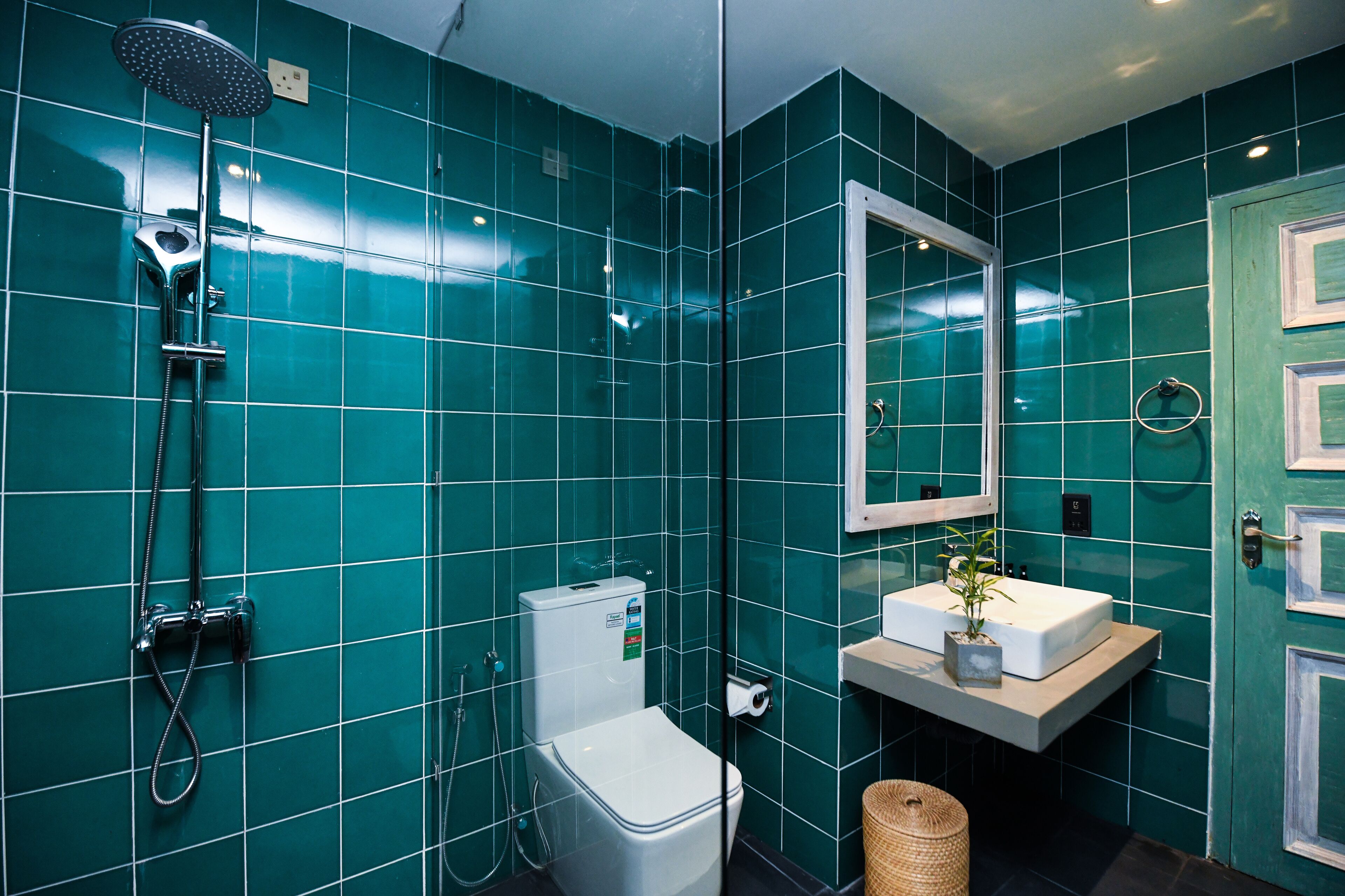A bathroom with teal tiles, a white toilet, a shower with a handheld showerhead, a sink with a mirror above it, and a door with a handle.